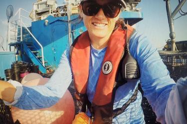 A woman wearing sunglasses, a radio, and a life vest on the deck of a vessel.