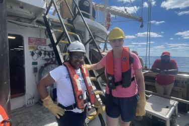 Three individuals stand smiling on the deck of a boat wearing orange vests, hard hats, and gloves