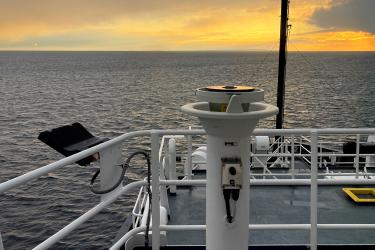 View of the setting sun from the bridge of a research vessel at sea. The yellow and gold sky peeks through low lying clouds that are in grays, purples, pinks, and apricots.