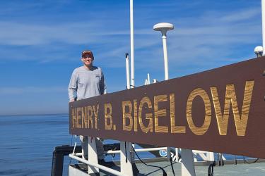 Sam Truesdell wears a baseball hat and a gray long sleeve shirt while standing on an upper deck of a research vessel at sea. He is standing behind a wooden sign “Henry B. Bigelow,” the ship’s name.