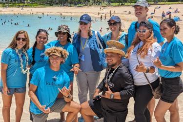 A group of 10 volunteers and staff in matching shirts pose for a photo on a beach