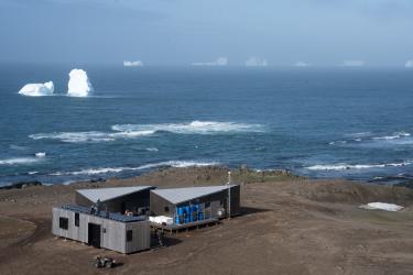 Three new camp buildings sit in the lower left corner of a dirt plane. In the background is the ocean and several large icebergs. The sky is blue and hazy.