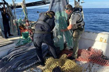 Two men in vests and khakis stand on a commercial fishing boat with nets at their feet. They are inspecting a net hanging between them.