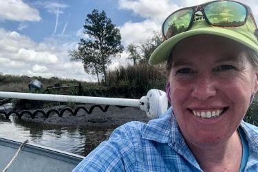 A person wearing a green baseball cap sits in a boat near a marsh