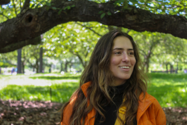 A young woman in an orange coat with long brown hair stands under a tree smiling in a green space
