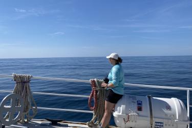 Person wearing a blue shirt and black shorts sitting on a boat looking out into the ocean, holding a camera. 