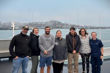 A group of people stand on the deck of a research vessel in front of a foggy, hilly, developed coastline