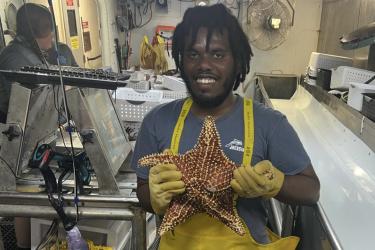 A young man wearing rubber gloves holding a rust colored sea star that is approximately 1 foot wide.