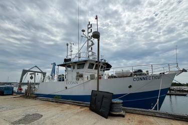 A research vessel is anchored by the dock. A frame is on the left side of the photo at the stern area of the boat. The bow area is to the right. The hull is painted with white and blue. Blue letters UCONN are painted on top of white paint on the side of the boat. Blue letters CONNECTICUT are painted on top of white paint on the hull of the bow area.