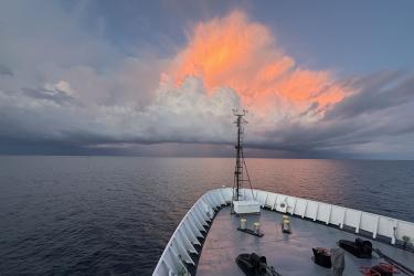 From the bow of a ship at sea, a burst of orange is seen in the sky above storm clouds and rain at the horizon.