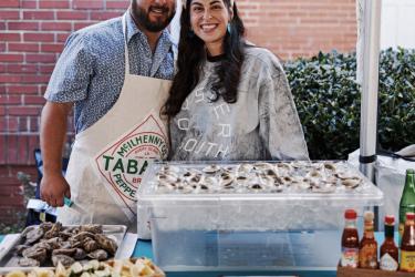 A married couple stand arm in arm, smiling, in front of an oyster booth with shucked oysters and garnishes. 