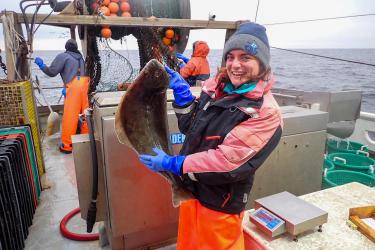 A fisheries observer wearing foul weather gear, a hat, a jacket, and blue rubber gloves stands on the back deck of a commercial fishing vessel holding an Atlantic halibut in her hands while posing for the camera.