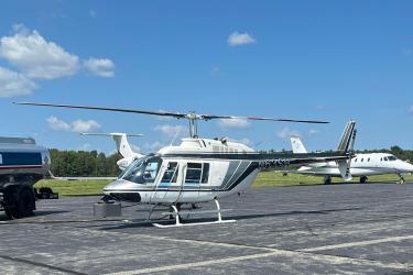 A mostly white helicopter in the center of the image on an airport runway. Small white passenger aircraft are parked in the background and there is a fuel truck to the left.