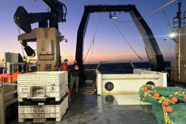 Two men stand at the back of a ship as the sky begins to lighten at the horizon with the approaching sunrise.
