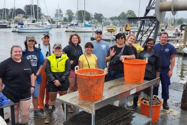 Group of eleven people smile on the back of a boat with three orange buckets filled with oysters.
