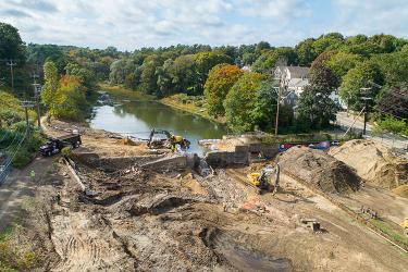 Aerial view of a construction site near the Holmes Dam with multiple excavators and dump trucks working on the land. A lush green landscape and residential buildings are visible in the background.