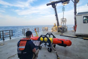 A scientist wearing a personal flotation device squats in front of a long-range autonomous underwater vehicle typing on a laptop while on the back deck of a research vessel at sea.