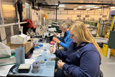 Two female scientists sit side by side at a lab bench holding pipettes.