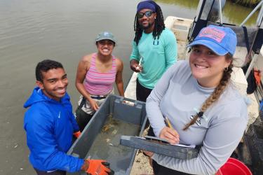 Two people standing in shallow water and two people on board a small boat smile at the camera. They are gathered around a bin used in a fish research project.