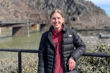 A young woman in a black puffy jacket stands against a railing overlooking a waterway crossed by an old bridge leading to the base of a mountain