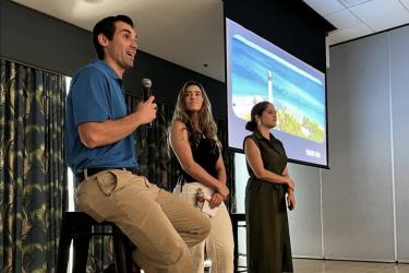 A man sitting on a stool speaks into a micophore while two women stand beside him.