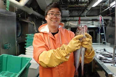 Nick is wearing a heavy orange rain coat and long yellow gloves. He holds up two pollock fish vertically, comparing their lengths to one another. We see more fish on a sorting table in the background.