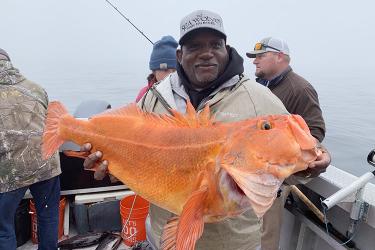 A bearded black man holds up a large, orange yelloweye rockfish using both hands and smiles for the camera. Three individuals stand behind him on a boat among fishing rods, buckets, caught fish, and other supplies.