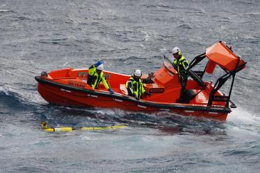 A small orange boat pulls up alongside a yellow glider floating at the ocean surface. There are three people in black and yellow safety clothing on the boat.
