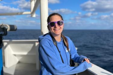 Kiersten stands at the taffrail of the ship and smiles for the camera. She is wearing a sweatshirt, purple sunglasses, and a pair of binoculars around her neck. the sky is blue with many clouds over a darker blue ocean.