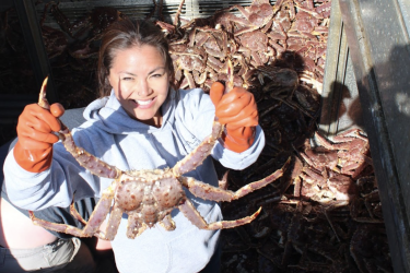 Mabel stands in a hold on a fishing vessel holding a red king crab by its legs.