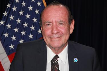 A White man in a suit and tie with a NOAA pin smiles in front of an American flag and black background.