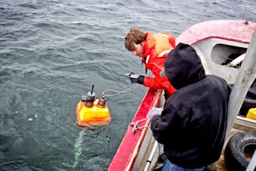 Two people stand on a boat and look out at a device on the water to conduct beluga whale acoustic monitoring.