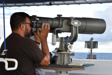 A man looks out of a large ship telescope over the water. 
