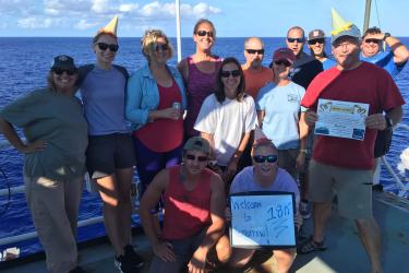 Group of people standing on a ship deck, wearing party hats and holding signs. 