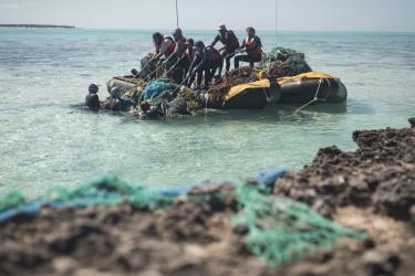 Two inflatable skiffs with many people on them, pulling up a large pile of derelict fishing gear out of the water. 