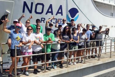 A group of people on the deck of a ship throwing shakas to the camera. 