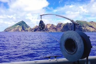 Close-up of a large reel and pulley on a ship, with blue water and jagged mountains in the background. 