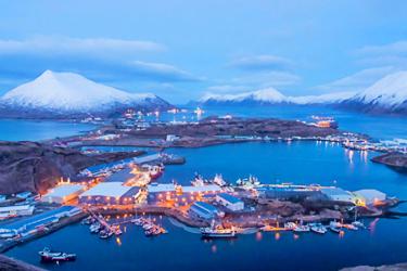 Aerial photograph of Dutch Harbor, Alaska. Snowcapped mountains surrounded by water and the lit up harbor town during the blue hour reflect creating a gradient between sea and sky. Credit: NOAA Fisheries