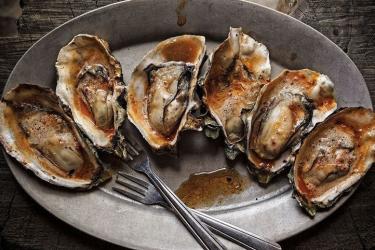 Six grilled Hog Island oysters drizzled in barbecue chipotle butter, on a serving dish with two small forks.