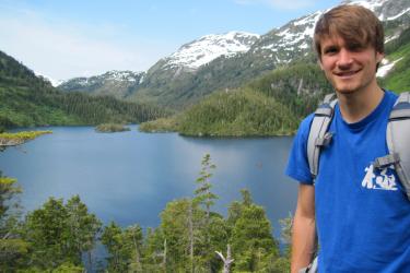 Brunette man wearing blue shirt and bookbag hiking near a lake with mountains in the distance
