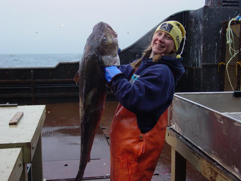 Photo of Ingrid Spies holding up a large Pacific cod on a boat deck.