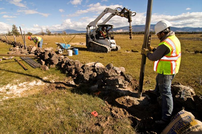 Two people in construction hats and vests working in a field in front of a piece of heavy machinery