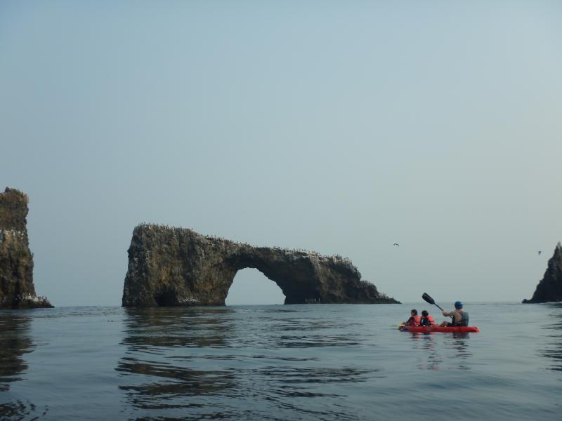 Three people in a red kayak paddle near a large stone arch.