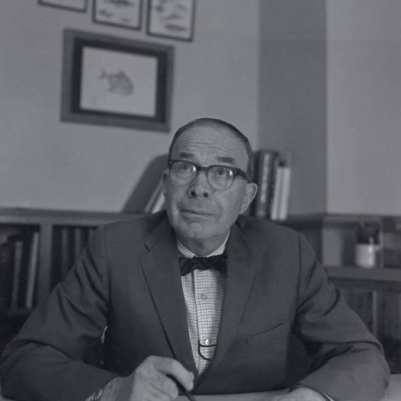 A black and whote photograph of a middle aged man at a desk with half-height bookcases filled with books in the background. The man wears glasses, a sport jacket with a light plaid shirt and dark, slender bow tie. 