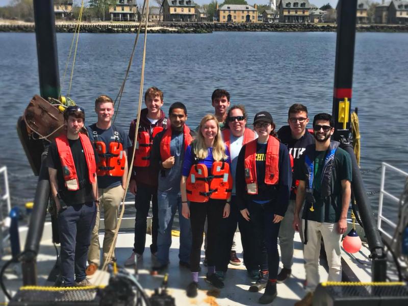 Ten people wearing orange life vests stand at the back end of a research vessel with a large, metal, black arch above them. The vessel is on the water with several buildings on the shore in the background. 