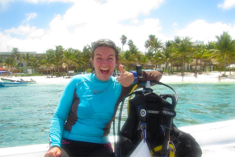 Catie Alves smiles and gives the thumbs up as she poses next to her dive gear while sitting on a boat. She is wearing a light blue long-sleeved shirt and black leggings. There is blue water and a beach with palm trees in the background.