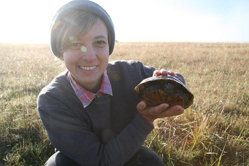 Tori Kentner squats in a grassy field wearing a gray beanie hat and sweater while holding a western pond turtle.