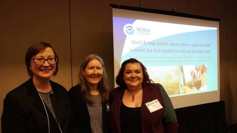 Three female scientists stand in front of a slide presentation in a conference room. The slide reads “Shelf & bag oyster aquaculture cages provide habitat for fish similar to natural boulder reefs”. 