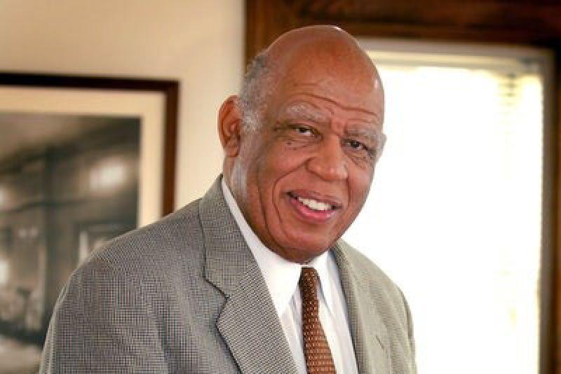 Color image of a bald African American man wearing a light colored suit and shirt, with a dark tie. He is standing in front of a window in an office setting, smiling at the camera.