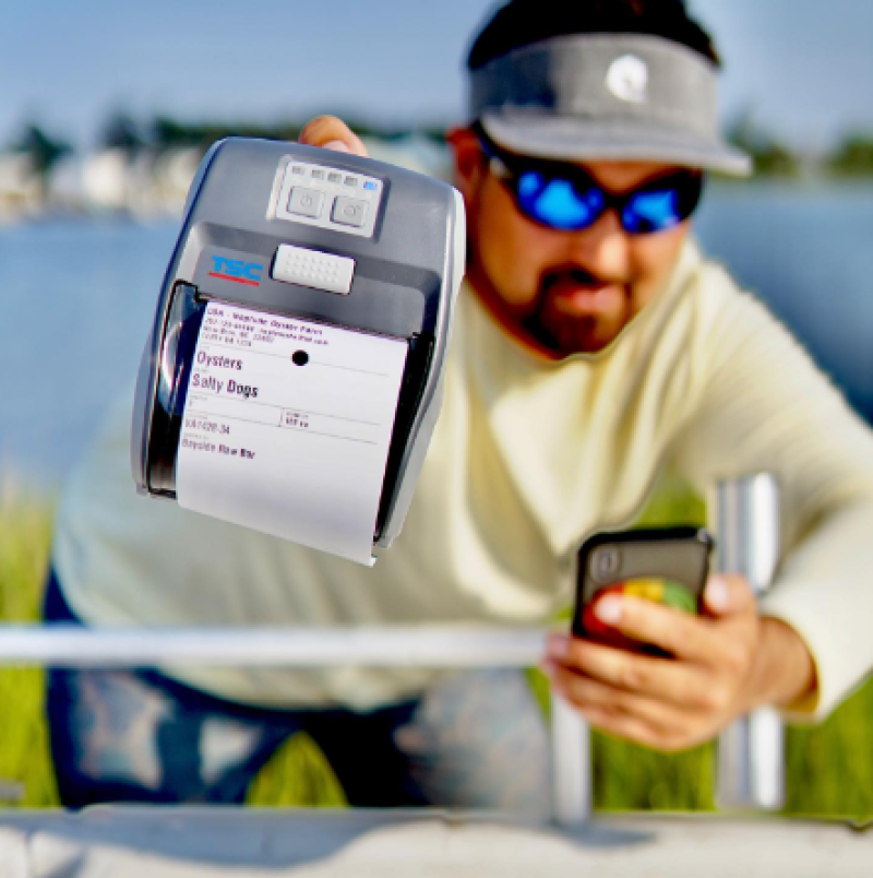 A shellfish grower holding a BlueTrace printer and oyster label in one hand, and a smartphone in the other hand.